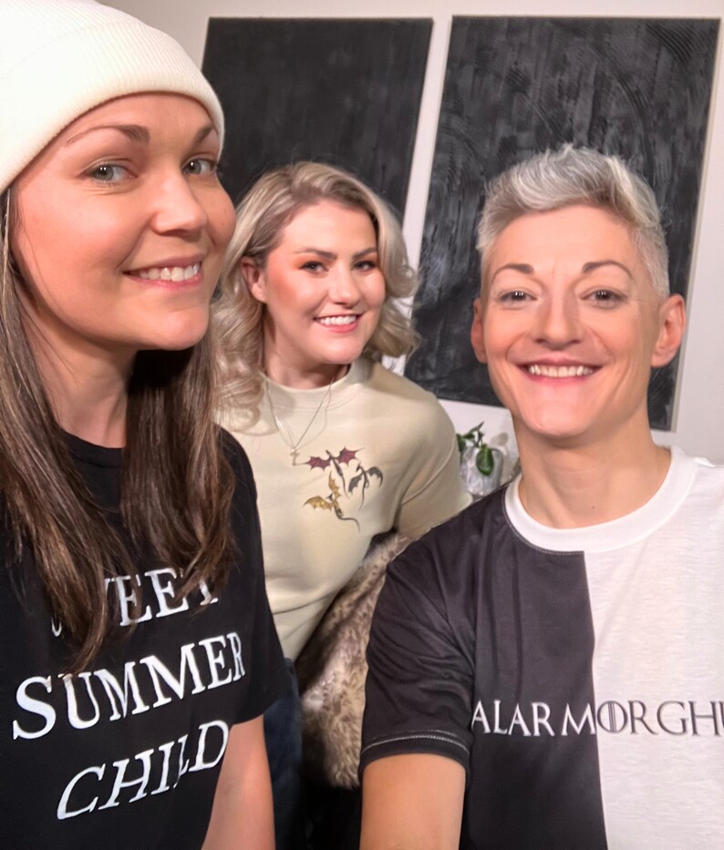 May include: Three people are wearing t-shirts with different designs. The person on the left is wearing a black t-shirt with the text "Sweet Summer Child". The person in the middle is wearing a light beige t-shirt with a design of three dragons. The person on the right is wearing a black and white t-shirt with the text "Alar Morghi".