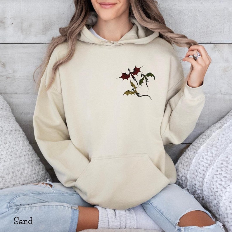 May include: A sand-colored hoodie with a pocket featuring a graphic of three dragons flying in formation. The dragons are red, green, and black.