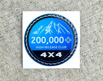 Op de afbeelding: Een ronde sticker met een blauwe gradiëntachtergrond en een witte bergafbeelding. De tekst luidt 200,000+ HIGH MILEAGE CLUB in het wit, met 4X4 in het wit op een zwarte horizontale balk onderaan. De rand heeft een zwart-wit bandenprofielpatroon.
