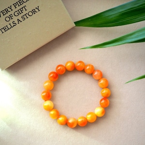 May include: A bracelet made of orange and yellow beads. The bracelet is on a light brown surface with a green leaf in the background.