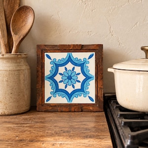 May include: A square ceramic tile featuring a blue and white floral pattern with symmetrical scrollwork, encased in a dark brown wooden frame. The tile displays a central flower motif surrounded by intricate blue geometric shapes.