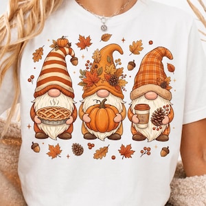 Può includere: Una t-shirt bianca con la grafica di tre gnomi con cappelli a tema autunnale. Lo gnomo a sinistra tiene una torta, quello centrale una zucca e quello a destra un bicchiere di caffè con la scritta FALL VIBES, circondati da foglie e ghiande.