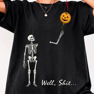 May include: A black t-shirt featuring a white skeleton graphic with one detached arm holding an orange jack-o'-lantern balloon. The text "Well, Shit..." is printed in white script below the skeleton. The design is centered on the front of the shirt.