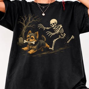 May include: A black t-shirt featuring a graphic print of a small brown and black Yorkshire Terrier running with a bone in its mouth, pursued by a human skeleton. In the background, there is a bare tree and a tombstone with the letters RIP.