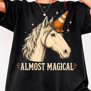 Puede incluir: Una camiseta negra con una ilustración gráfica de un caballo que lleva un cono de tráfico naranja en la cabeza como si fuera un cuerno, sujeto con una cuerda. El diseño incluye estrellas y el texto ALMOST MAGICAL impreso en letras claras.