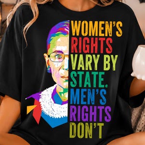 May include: A black t-shirt featuring a colorful, geometric pop-art style portrait of Ruth Bader Ginsburg on the left side. The right side displays the text: WOMEN'S RIGHTS VARY BY STATE. MEN'S RIGHTS DON'T, with each word in a different vibrant color.
