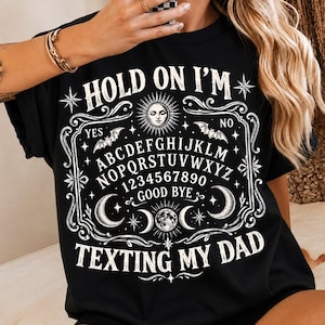 Könnte beinhalten: Ein schwarzes T-Shirt mit einem weißen Grafikdruck eines Spirit-Board-Designs. Der Text lautet "HOLD ON I'M TEXTING MY DAD" und enthält Illustrationen von Sonne, Mondphasen, Sternen und Fledermäusen um das Alphabet und die Zahlen 0 bis 9.