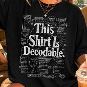 Op de afbeelding: Een zwart t-shirt met witte tekst en educatieve diagrammen over fonetiek en leesregels. De centrale tekst luidt This Shirt Is Decodable, omringd door kleinere vakken met details over gesloten lettergrepen, tweeklanken en leesstrategieën.