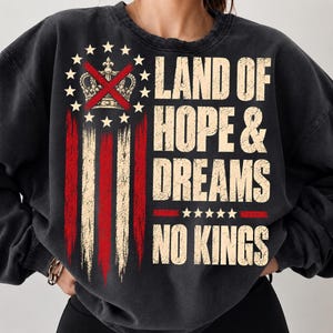 May include: A charcoal black crewneck sweatshirt featuring a distressed graphic print. The design includes a circle of white stars surrounding a crown crossed out with red lines, vertical red and white stripes, and the text LAND OF HOPE & DREAMS NO KINGS.
