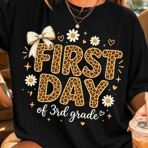 May include: A black t-shirt featuring the text FIRST DAY of 3rd grade in a leopard print pattern. The design includes a large cream-colored bow with black dots, white daisy flowers, small yellow hearts, and four-pointed stars surrounding the text.
