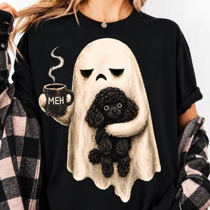 Puede incluir: Una camiseta negra con un gráfico de un fantasma beige que sostiene una taza negra con el texto MEH y abraza a un pequeño caniche negro. El fantasma tiene una expresión neutral y sale vapor de la taza. Se lleva con una camisa de franela.