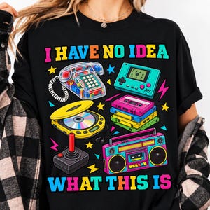 Pode incluir: Uma t-shirt preta com o texto I HAVE NO IDEA WHAT THIS IS em cores vivas. O gráfico inclui um telefone transparente, uma consola de jogos portátil, uma pilha de cassetes, um leitor de CD portátil, um joystick e um boombox.