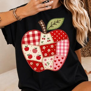 Könnte beinhalten: Ein schwarzes T-Shirt mit einer großen, zentralen Apfelgrafik, die aus verschiedenen Patchwork-Segmenten besteht. Das Apfeldesign enthält Muster aus rot-weißem Vichy-Karo, roten Punkten, kleinen Äpfeln, weißen Blumen und roten Herzen.