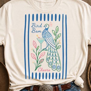 May include: A cream-colored t-shirt featuring a graphic of a blue peacock standing next to a martini glass. The design includes pink and green foliage, blue vertical stripes on the sides, and the text "Bird Bam" and "Cheers" in blue and pink.