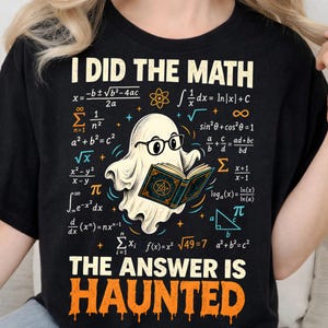 May include: A black t-shirt featuring a graphic of a white ghost wearing black glasses and reading a book. The text reads I DID THE MATH and THE ANSWER IS HAUNTED, surrounded by various mathematical formulas and symbols in orange, blue, and white.
