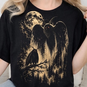 Puede incluir: Una camiseta negra con un estampado gráfico en tono dorado de una criatura grande similar a una polilla con ojos rojos brillantes, una polilla más pequeña en una rama, una luna llena y un paisaje forestal. Diseño con detalles de tinta texturizada.