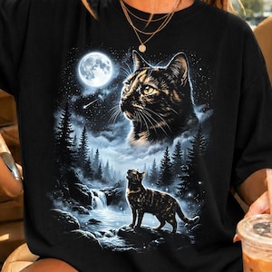 May include: A black t-shirt featuring a graphic print of a tortoiseshell cat looking up at a full moon in a starry night sky. Below, a smaller tortoiseshell cat stands on a rock near a waterfall and a dark forest of pine trees under the moonlight.