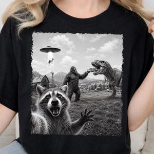 Puede incluir: Una camiseta negra con un estampado gráfico en blanco y negro de un mapache tomándose un selfie en primer plano. Al fondo, un Bigfoot le da una porción de pizza a un T-Rex, mientras un OVNI abduce a una vaca cerca de un granero rústico.