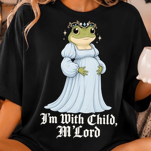 Op de afbeelding: Een zwart t-shirt met een afbeelding van een groene kikker in een lichtblauwe jurk met empire-taille en een kroon met een blauwe edelsteen. De kikker houdt de handen op de buik. Onder de afbeelding staat de tekst: I'm With Child, M'Lord.