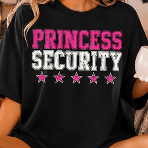 May include: A black short-sleeved t-shirt featuring the text PRINCESS in pink and SECURITY in white, both in a distressed block font. Below the text are five pink stars arranged in a horizontal row across the center of the chest.
