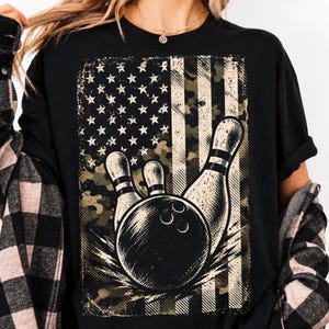 May include: A black t-shirt featuring a graphic print of a bowling ball striking pins. The design incorporates a distressed American flag pattern with a camouflage overlay in shades of green, brown, and tan, set against a rectangular background.