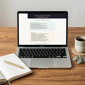 Puede incluir: Un portátil que muestra un diario titulado "Perimenopause Mood & Symptom Journal" está sobre un escritorio de madera, junto a un cuaderno abierto y un bolígrafo dorado. Una taza de café descansa sobre un posavasos a la derecha.