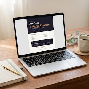 May include: A laptop displaying an "Anxiety Trigger Tracker" document, with text in dark blue and gold. A notebook with a gold pen, and a mug of coffee on a coaster are on a wooden table. The laptop is open, showing the document's title and content.