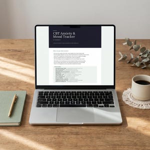 May include: A laptop displaying a "CBT Anxiety & Mood Tracker" document on a wooden desk. Beside the laptop are a pen, a notebook, and a cup of coffee on a coaster. The screen shows text about tracking mood and anxiety.