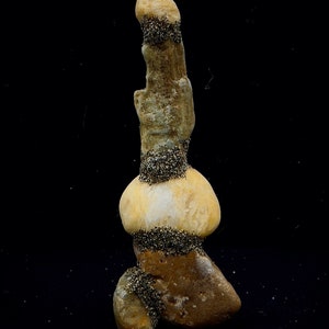 May include: A tall, stacked rock formation with a smooth, light brown surface. The rock is covered in small, black specks, creating a textured look. The rock is isolated against a black background.
