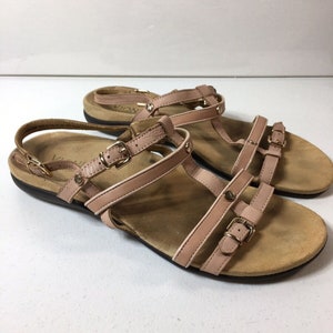 以下が含まれることがあります： 複数のストラップとバックルが付いた薄いピンクの革のサンダル。サンダルはコルクのフットベッドと茶色のアウトソールを持っています。