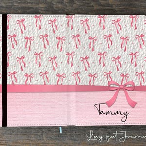 Op de afbeelding: Een roze notitieboek met een strikdesign. De bovenste helft van het notitieboek heeft een witte achtergrond met een patroon van roze strikken. De onderste helft is effen roze met de naam "Tammy" en de tekst "Lay Flat Journal".