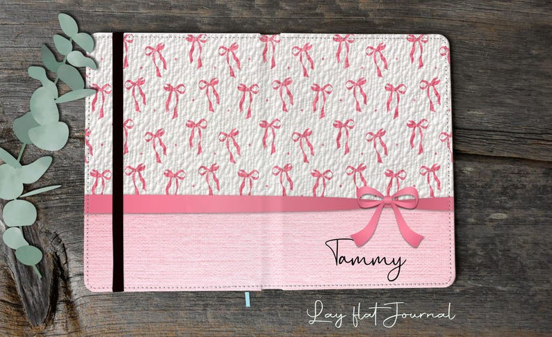 Op de afbeelding: Een roze notitieboek met een strikdesign. De bovenste helft van het notitieboek heeft een witte achtergrond met een patroon van roze strikken. De onderste helft is effen roze met de naam "Tammy" en de tekst "Lay Flat Journal".