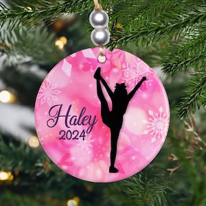 May include: A round, pink Christmas ornament with a gymnast silhouette and the name "Haley" and the year "2024" in a script font. The ornament has a snowflake design and is hanging from a string with silver beads.