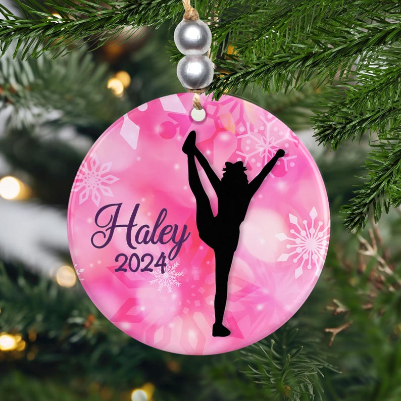 May include: A round, pink Christmas ornament with a gymnast silhouette and the name "Haley" and the year "2024" in a script font. The ornament has a snowflake design and is hanging from a string with silver beads.