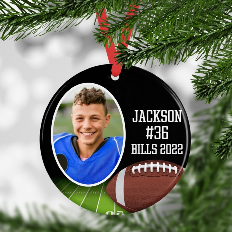 May include: A black, round Christmas ornament with a photo of a smiling person in a blue football uniform. The ornament has the text "JACKSON #36 BILLS 2022" and a football graphic. A red ribbon is attached.