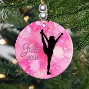 May include: Pink round Christmas ornament with a silhouette of a gymnast in a split. The ornament has the name "Julie" and the year "2024" in a script font, with snowflake designs.