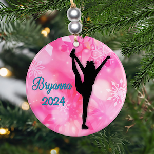 May include: A pink ceramic Christmas ornament with a gymnast silhouette. The ornament has a pink background with white snowflakes and the name "Bryanna" in teal script, along with the year "2024". The ornament is hanging from a string with silver beads.
