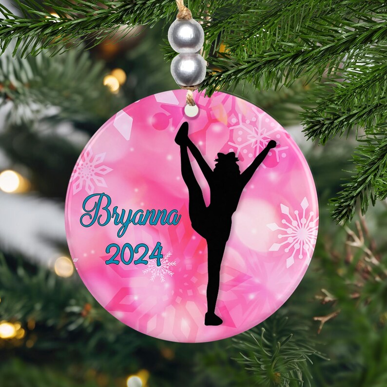 May include: A pink ceramic Christmas ornament with a gymnast silhouette. The ornament has a pink background with white snowflakes and the name "Bryanna" in teal script, along with the year "2024". The ornament is hanging from a string with silver beads.