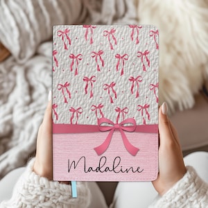 Op de afbeelding: Een gepersonaliseerd notitieboek met een roze strik ontwerp. De bovenste helft van het notitieboek heeft een witte achtergrond met een herhaald patroon van roze strikken. De onderste helft is roze met de naam "Madaline" in zwart.