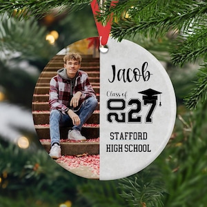 May include: A round ceramic ornament featuring a photo of a person in a plaid shirt and jeans sitting on stairs on the left side, and white text on the right reading Jacob, Class of 2027, and Stafford High School with a black graduation cap icon.