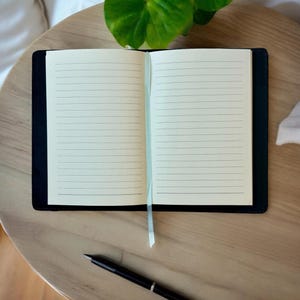 Op de afbeelding: Een open notitieboek met gelinieerde pagina's en een zwarte kaft, met een lichtblauw lint als bladwijzer. Een zwarte pen ligt op een lichtbruin houten oppervlak naast het notitieboek. Een groene plant is zichtbaar op de achtergrond.