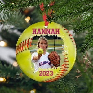 May include: A yellow softball ornament with red stitching and a white, cracked photo frame. The photo shows a girl holding a softball and a glove. The ornament has the name "HANNAH" and the year "2023" printed on it.