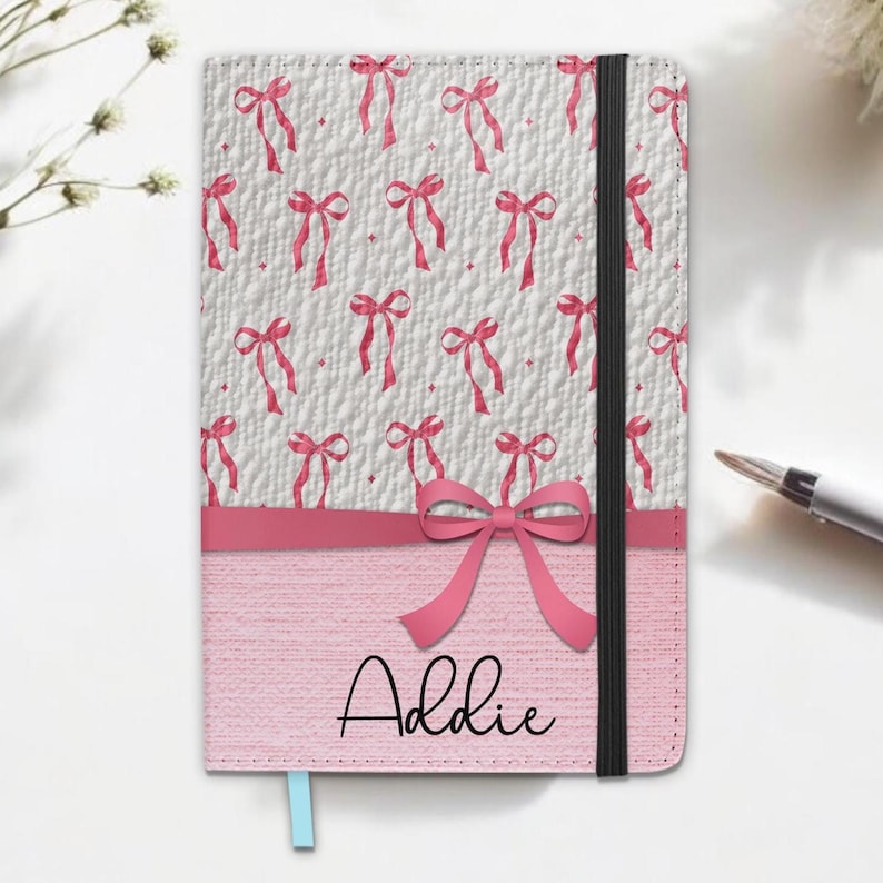 May include: A personalized notebook with a pink bow design. The top half features a white background with a repeating pattern of pink bows. The bottom half is pink with the name "Addie" in cursive. A black elastic band and a blue ribbon bookmark are also visible.