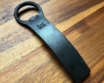 May include: A black metal bottle opener with a rounded opening and a curved handle. The opener has the letter "M" engraved on the handle.