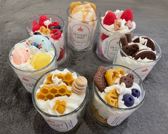 May include: A collection of dessert-themed candles in clear glass cups. Each candle is topped with decorative wax elements resembling various desserts, including fruit, cookies, and whipped cream. The candles have the text "Dessert Light" on the label.