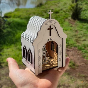 May include: A small, laser-cut wooden model of a church with white walls and dark brown edges. The structure features a cross on the roof, arched windows, and an open front entrance displaying a decorative cutout of a religious figure inside.