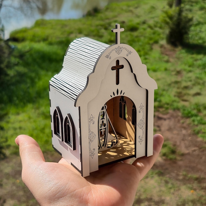 May include: A small, laser-cut wooden model of a church with white walls and dark brown edges. The structure features a cross on the roof, arched windows, and an open front entrance displaying a decorative cutout of a religious figure inside.