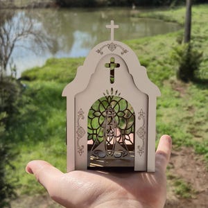 May include: A small, light-colored wooden structure shaped like a church, featuring an arched entrance with a decorative lattice pattern and a cutout of a figure in the center. The top is adorned with a cross and intricate filigree engravings.