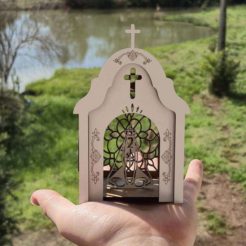 May include: A small, light-colored wooden structure shaped like a church, featuring an arched entrance with a decorative lattice pattern and a cutout of a figure in the center. The top is adorned with a cross and intricate filigree engravings.