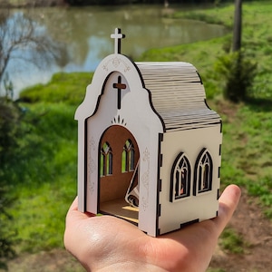 May include: A small, white, laser-cut wooden model of a church with dark brown edges. The structure features an arched entrance, a cross at the peak, and arched windows on the side. Intricate decorative patterns are etched onto the front facade.